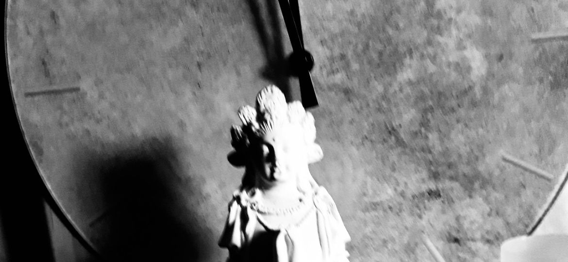 Statue mit gefalteten Händen vor einer Uhr mit Zeigern.